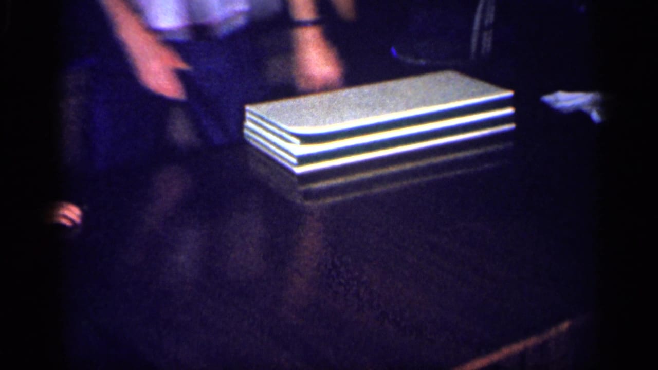 A stack of white rectangular panels rests on a dark tabletop beside partially visible adults.