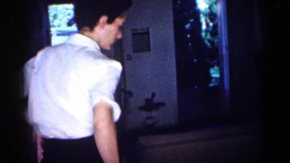 A man in a white shirt stands near a doorway or entry area in a dim 1959 Michigan home-movie interior.