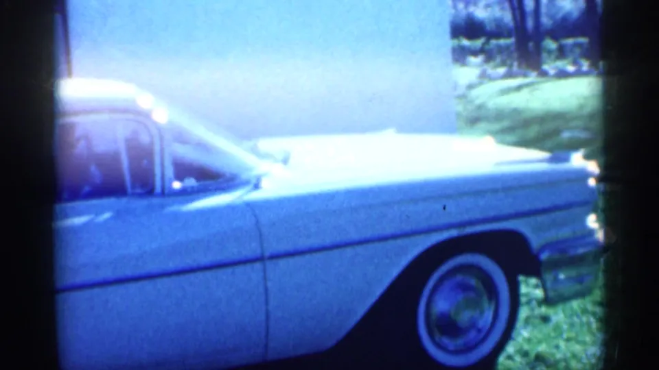 A white mid-century car appears outside near grass and trees in the 1959 Michigan home-movie footage.