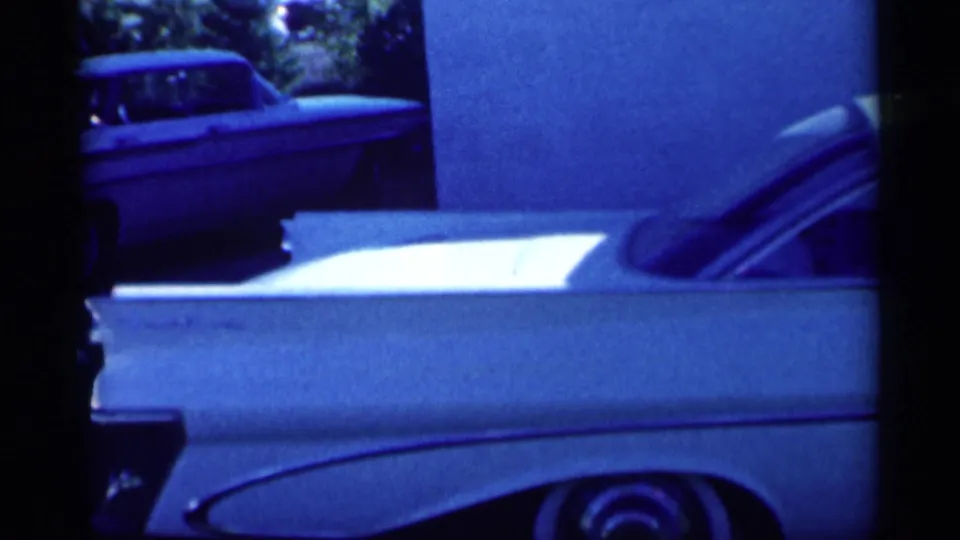 Blue-tinted home-movie frame of the white sedan's rear fin and trunk area, with another parked car partly visible in the background.