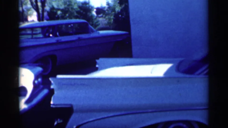 1959 Michigan home-movie frame showing a light station wagon and part of a white sedan near a green suburban yard.