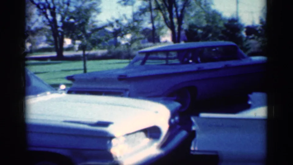 A 1959 Michigan suburban frame shows the station wagon behind the white sedan, with grass and trees beyond the driveway.