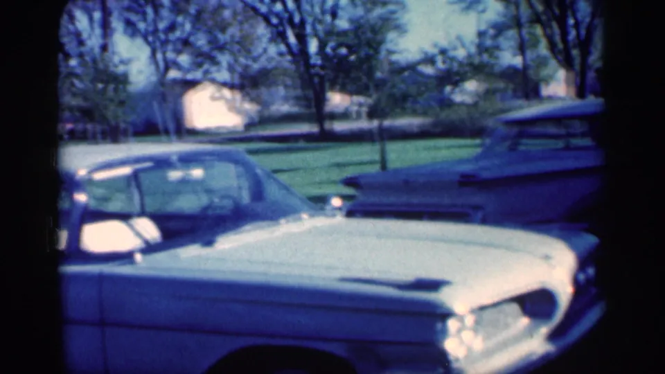 1959 Michigan home-movie frame with a white sedan foreground, a light station wagon, trees, and nearby houses in the background.