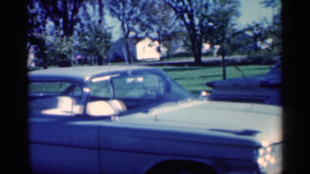 Light blue car seen close beside a parked car on a sunny suburban street.