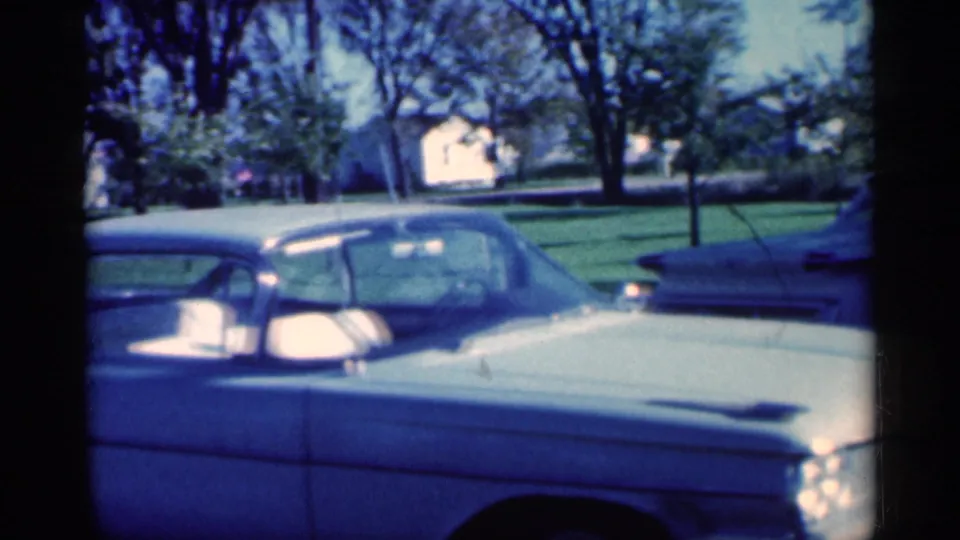 1959 Michigan 8mm frame showing a light blue car close to the camera with another parked car, lawns, trees, and houses beyond.
