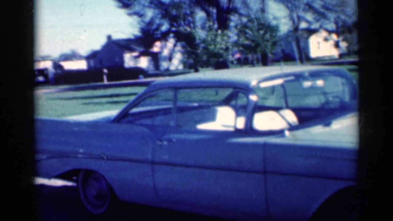 1959 Michigan home-movie frame with a pale blue sedan in side view, another car behind it, lawns, trees, and houses in the background.