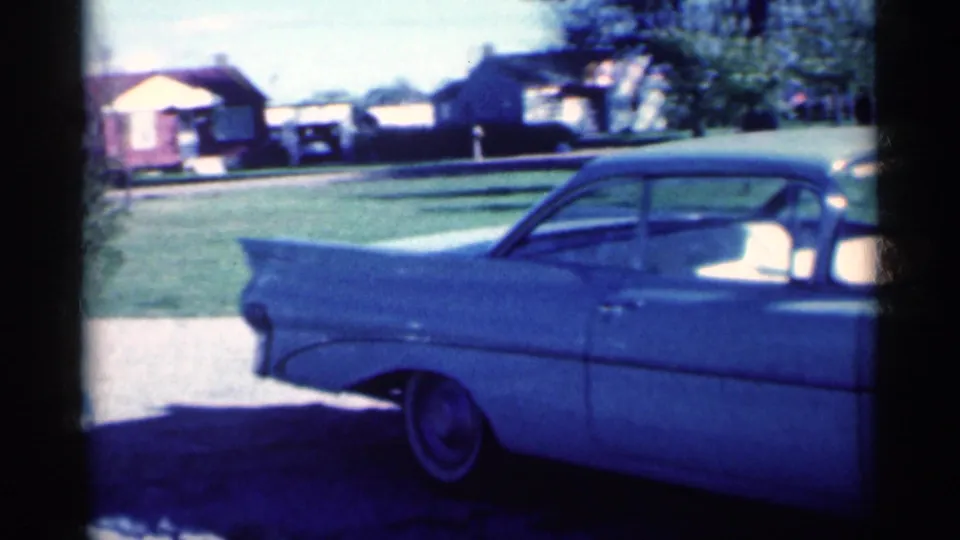1959 Michigan 8mm frame showing the rear side of a pale blue sedan with tail fin styling, curb, lawn, trees, and houses.