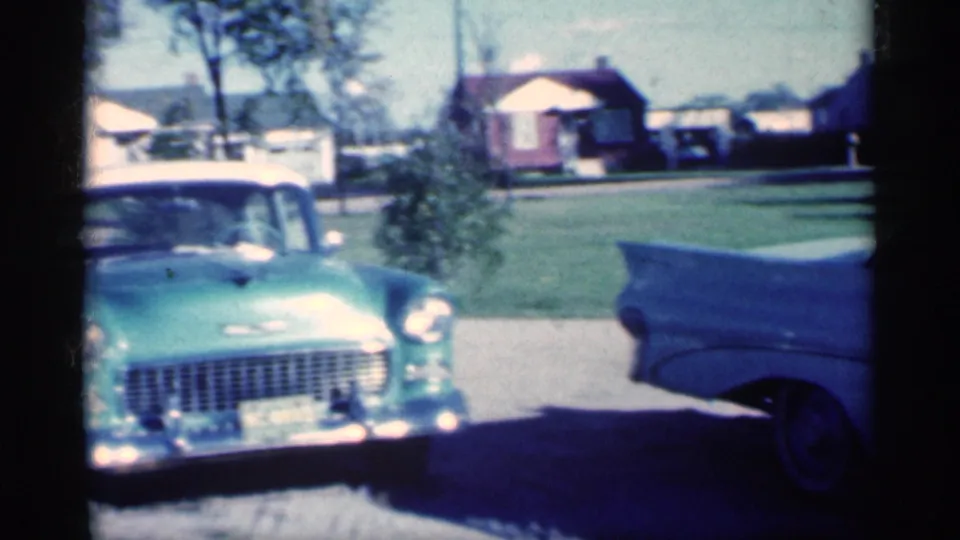 1959 Michigan frame showing a turquoise Chevrolet-style car with a white roof near a blue car, with lawn, trees, and houses behind.