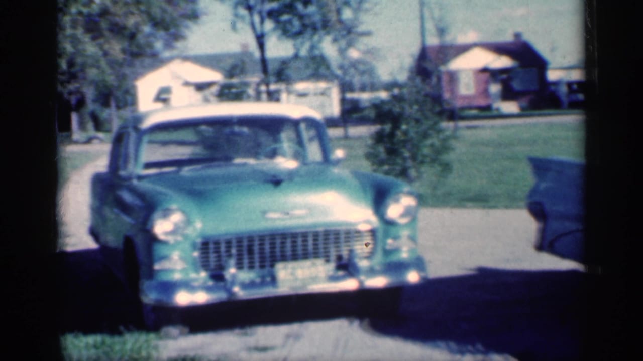 Green 1950s car parked near a suburban driveway with houses and trees behind it.