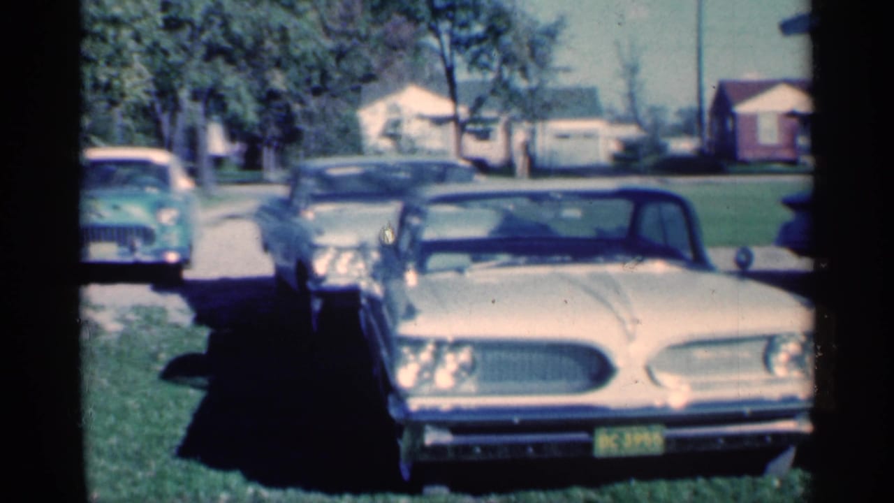 1959 Michigan frame with a cream-colored classic sedan in the foreground, another dark car beside it, and a green car near the street.
