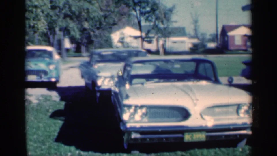 1959 Michigan frame with a cream-colored classic sedan in the foreground, another dark car beside it, and a green car near the street.