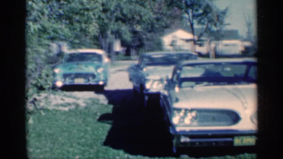 1959 Michigan home-movie view of a cream sedan close to camera, with a green car and darker parked automobile in the driveway area.
