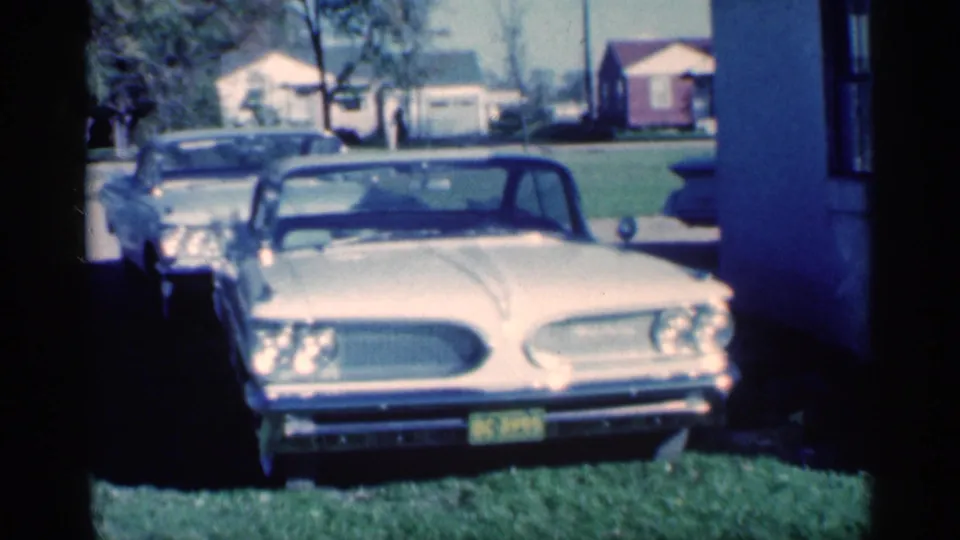 1959 Michigan frame showing a cream classic sedan facing camera, with a darker car behind it, lawn, house edge, and distant homes.
