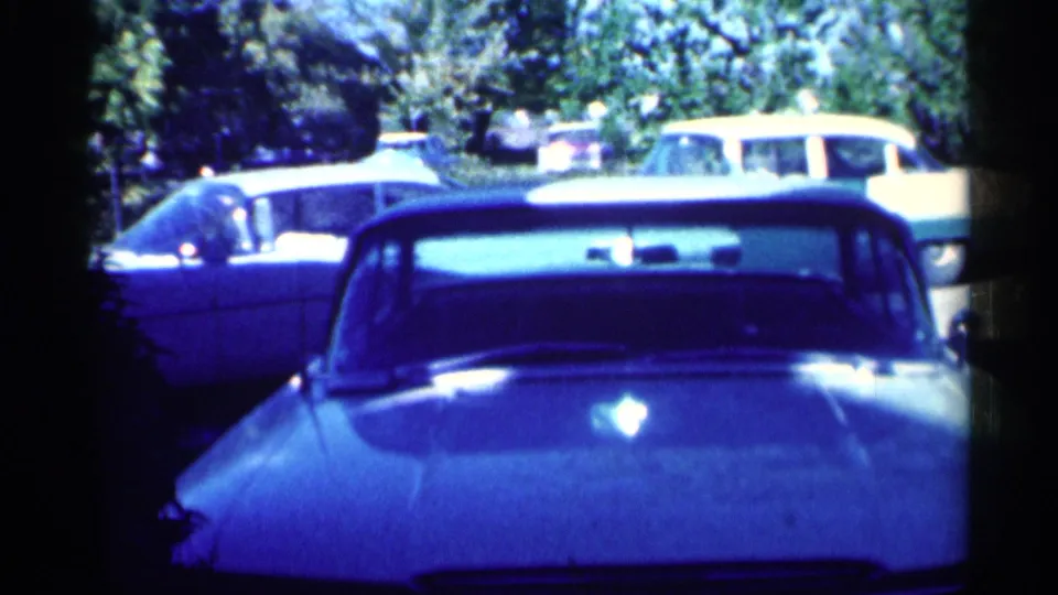 1959 Michigan 8mm frame showing a parked car in a suburban yard or driveway, with another car and trees in the background.
