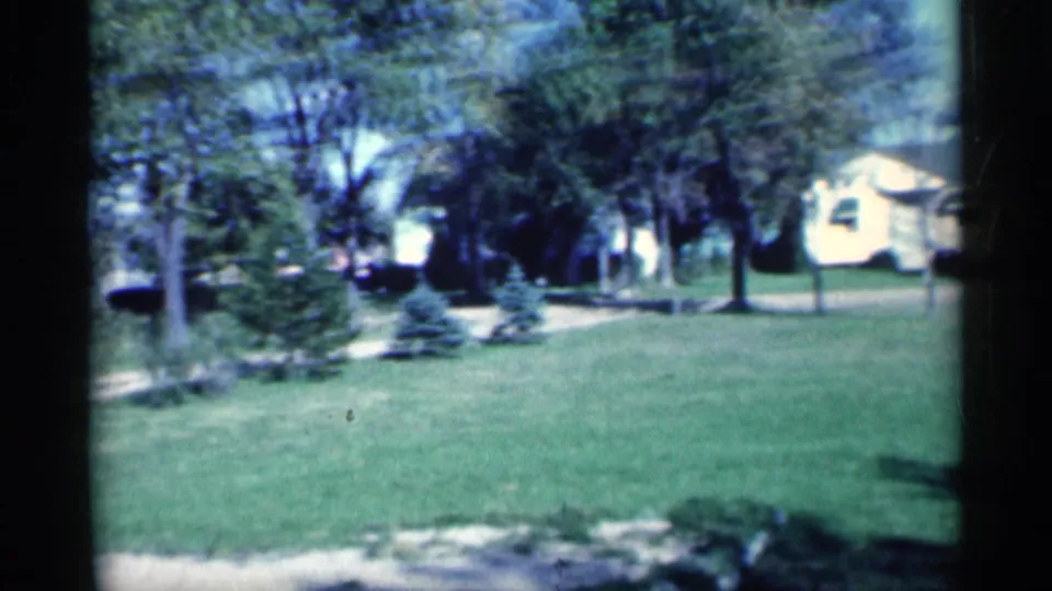 1959 Michigan home-movie frame showing a residential lawn with trees, shrubs, houses, and a chain-link fence.
