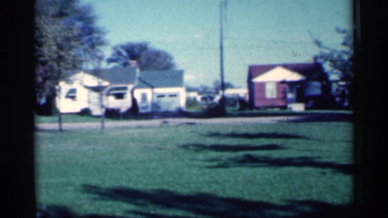 1959 Michigan 8mm view of single-story houses, open lawn, trees, driveway, and utility pole during a slow neighborhood pan.