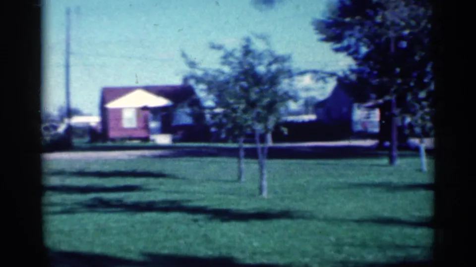 1959 Michigan 8mm frame with a red-sided house, pale gable, open lawn, young trees, and shaded residential frontage.