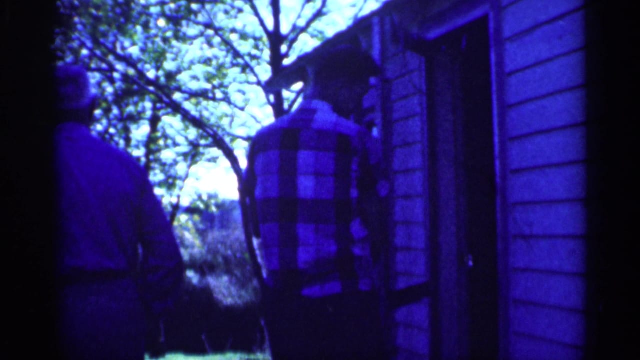Two men stand near the side of a wooden building, one wearing a plaid shirt.