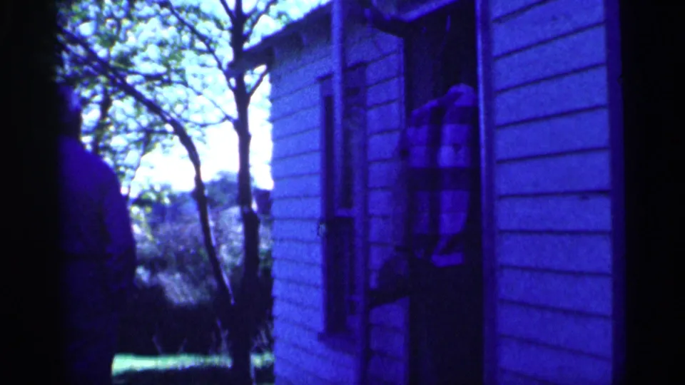 A plaid-shirted man appears to open or reach toward a screen door on a wooden-sided structure as another figure remains nearby.