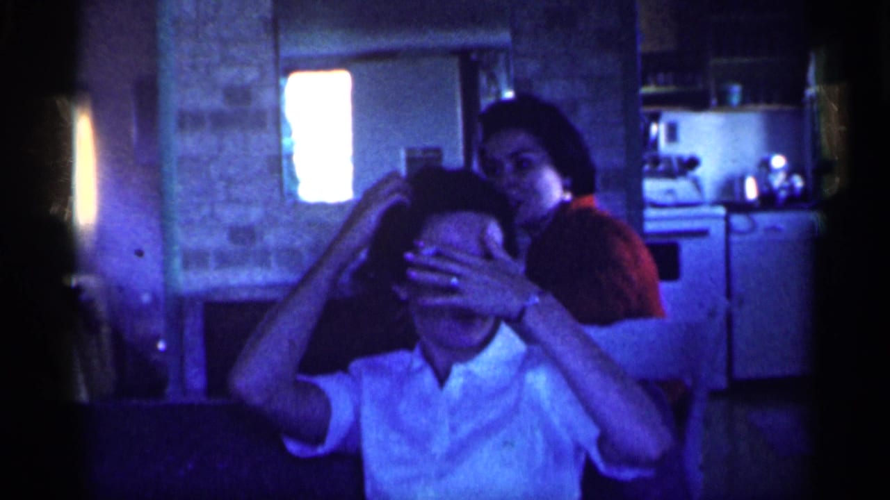 In a Michigan home interior from 1959, a woman in a white shirt shields her face as another woman in red stands behind her near a kitchen area.
