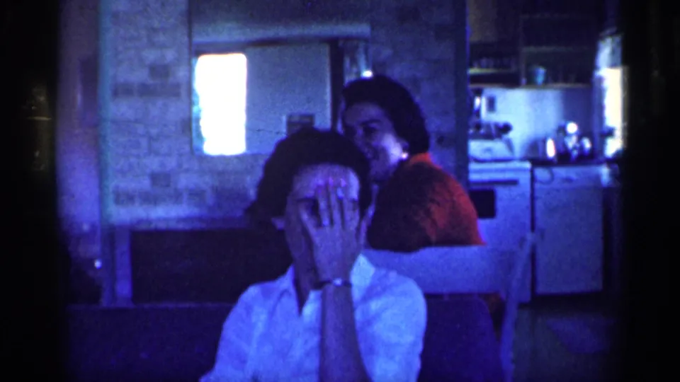The 1959 Michigan home-movie frame shows the woman in white covering her face while another woman in red smiles behind her near kitchen fixtures.