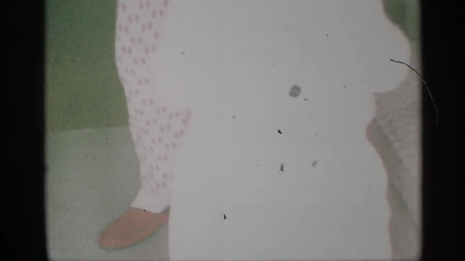 Blurry 1959 New York home-movie frame showing a young child in pale polka-dot clothing beside a green wall.
