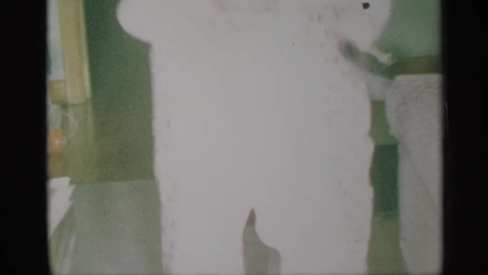 Blurry 1959 New York home-movie frame of a young child in pale polka-dot pajamas near a green interior wall.