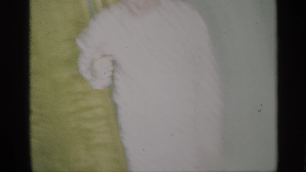 Overexposed home-movie frame showing part of a seated woman in a white dress beside green upholstery.