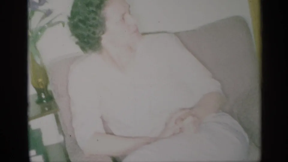 1959 New York home-movie frame showing a woman in a white dress seated in a tan armchair, with greenery behind her.