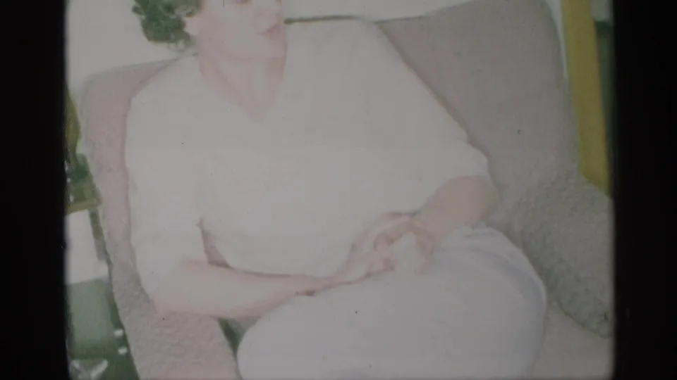 1959 New York 8mm frame of a woman seated indoors in a tan armchair, wearing a white dress with hands folded.