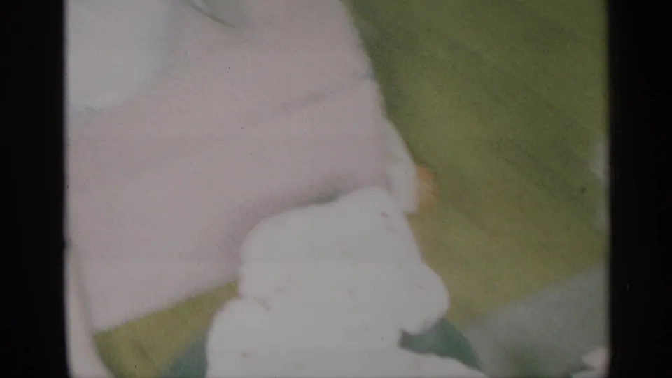 Cropped 1959 New York home-movie frame showing the woman's white dress, arm, and nearby green upholstery indoors.