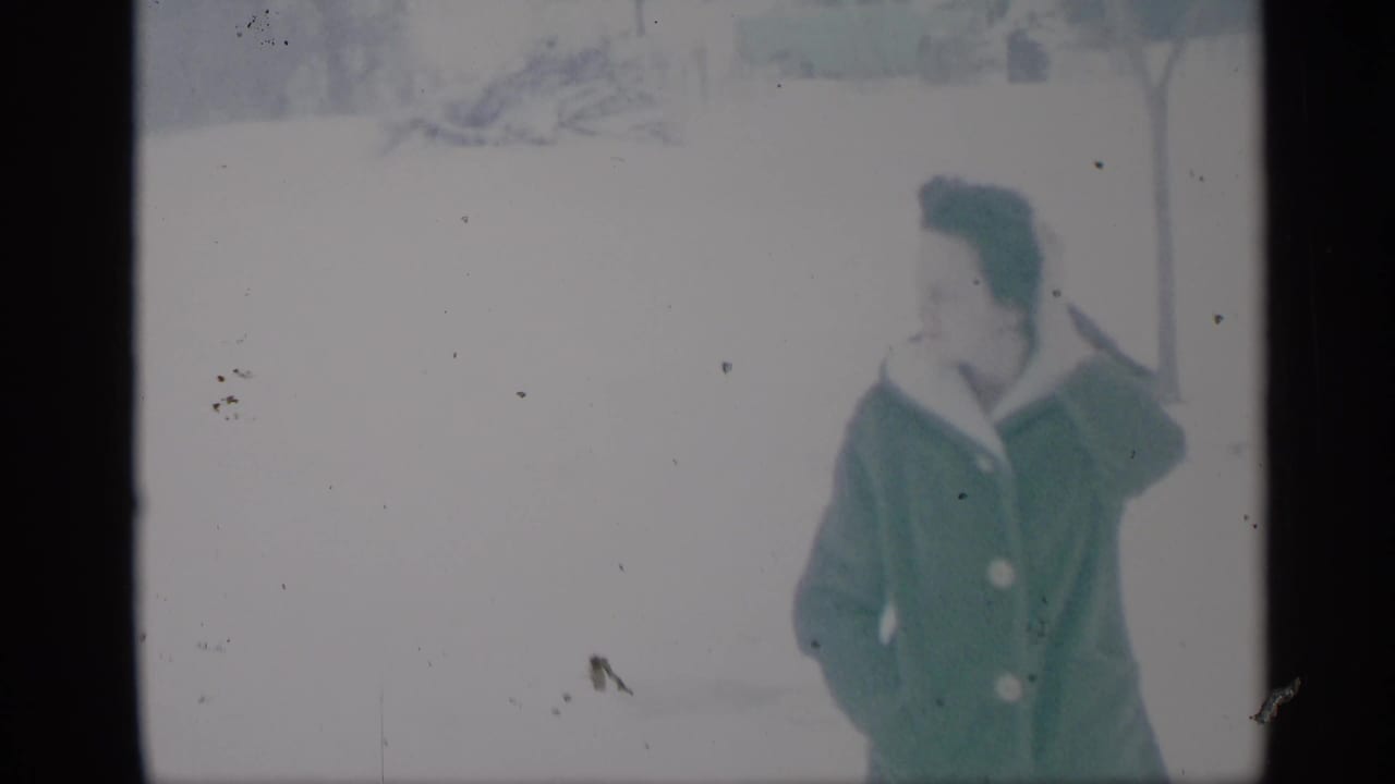 Woman in a green coat stands in snow with her hands near her coat.