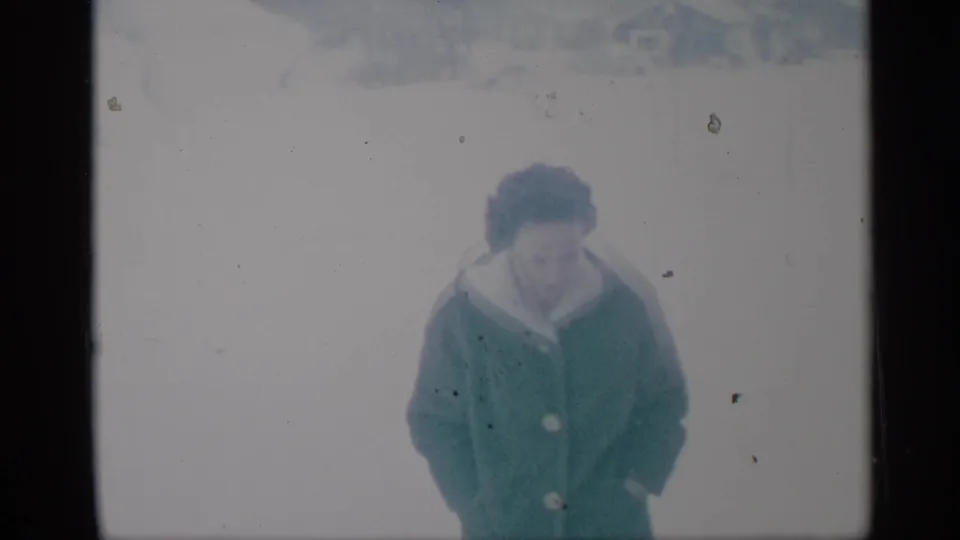 The woman walks closer through the snowy yard, her green coat prominent in the soft 8mm frame.