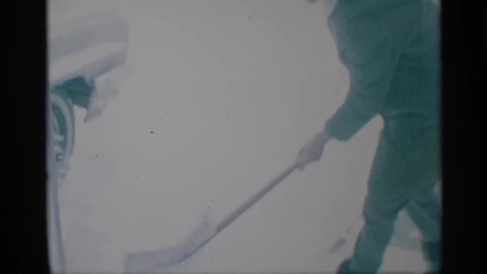 A woman in a green coat shovels snow near the edge of a parked car in a residential winter scene.