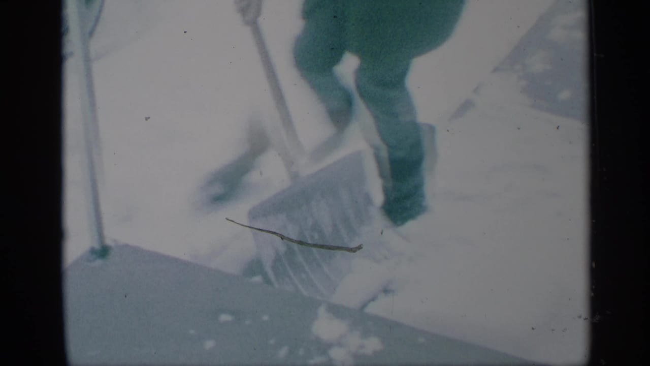 Person in winter clothing shoveling snow on pavement near a parked car.