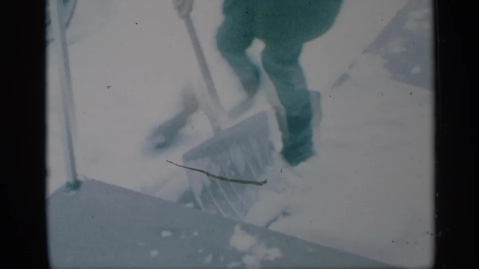 1959 New York home-movie frame showing a person in a green winter coat using a snow shovel on snowy pavement near a car.