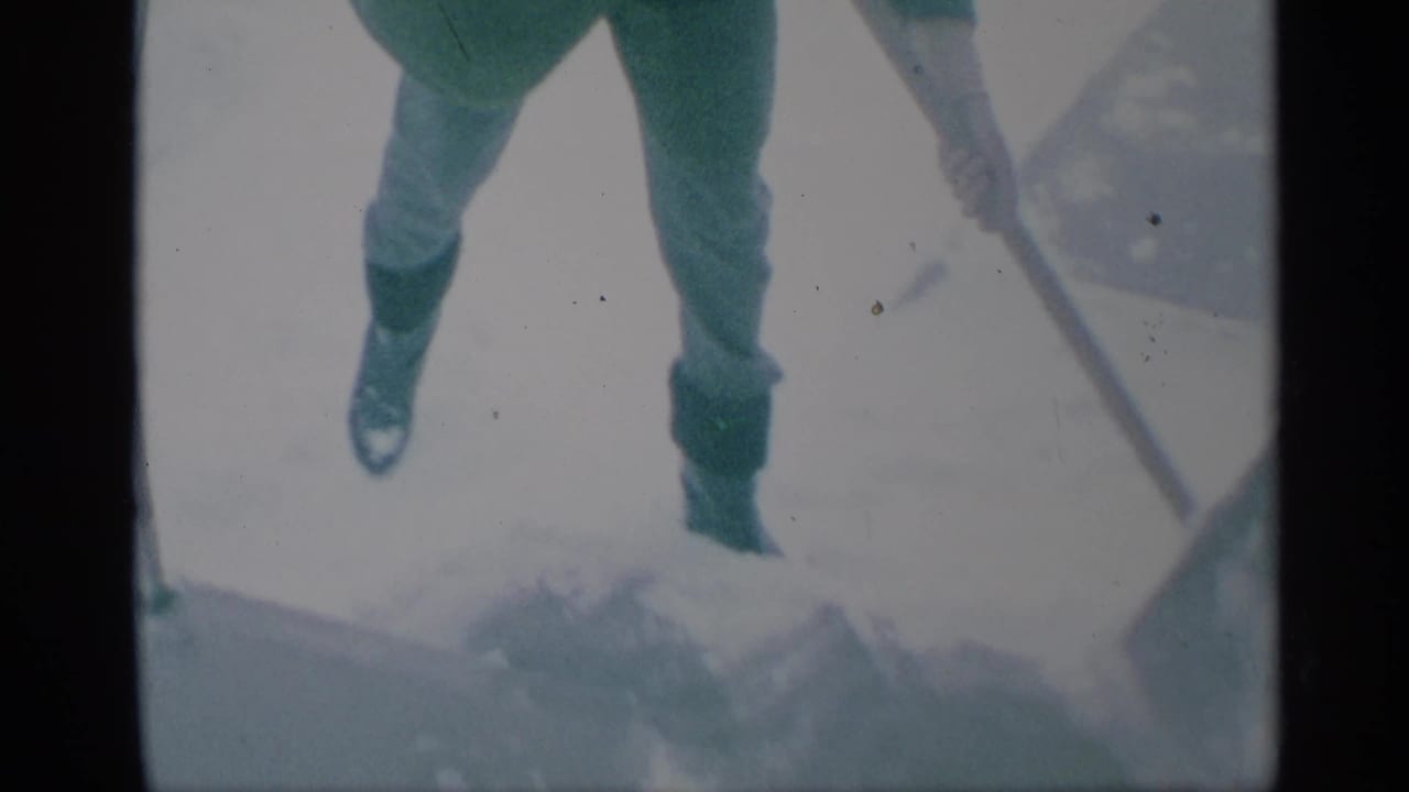 Lower body of a person standing on snow with a shovel nearby.