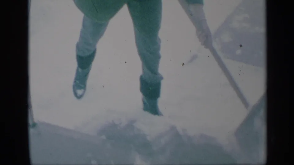 1959 New York frame with a person’s legs and boots on bright snowy pavement, with a shovel handle and snow buildup visible.