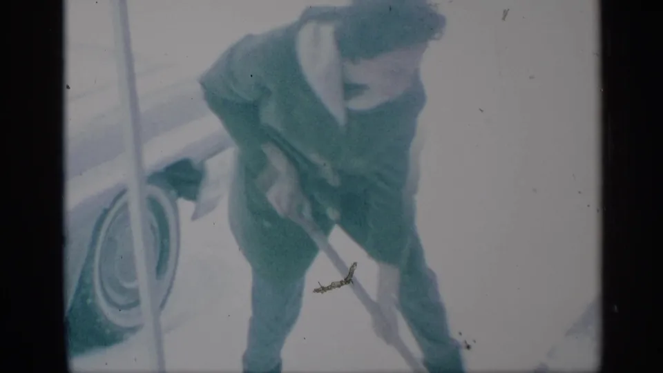 1959 New York home-movie frame showing a bundled person bending with a shovel beside snowy pavement and a partial car wheel.