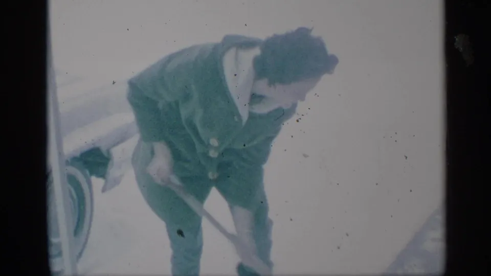 1959 New York frame showing a person in a green winter coat pushing or lifting snow with a shovel near a parked car.