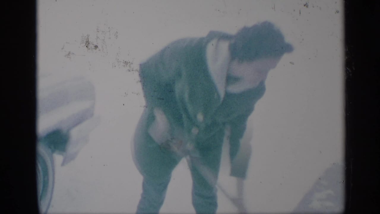 Woman in a dark coat bends forward while shoveling snow beside part of a vehicle.