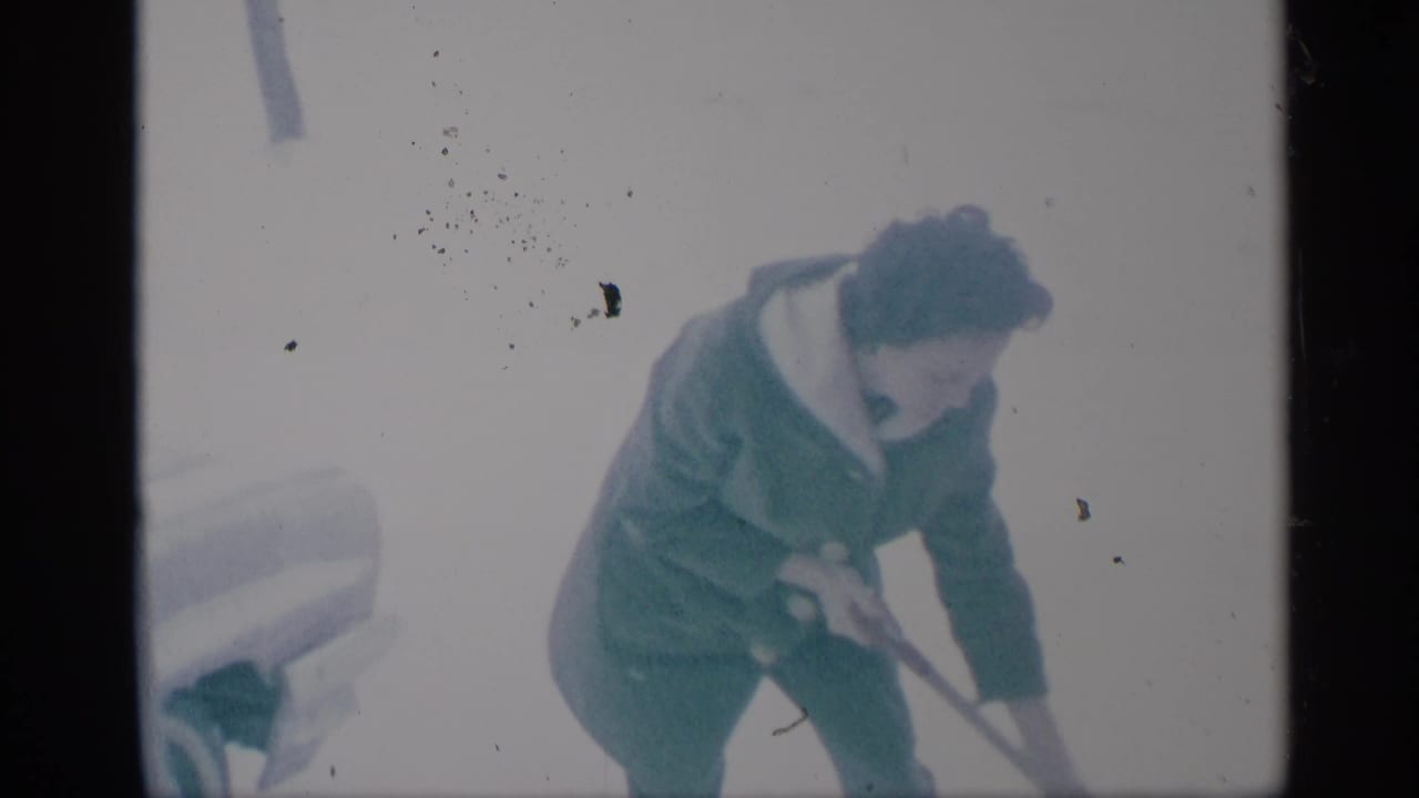 The woman continues shoveling snow in a soft, grainy 1959 New York home-movie frame with a vehicle partly visible at left.