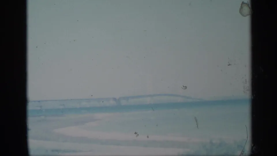 1959 New York 8mm frame showing a distant steel bridge across calm water under a pale, hazy sky.