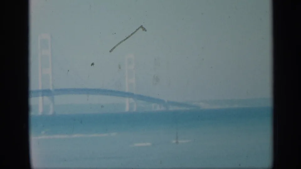 Faded 8mm frame with part of a suspension bridge and tower over water, marked by scratches and dust.