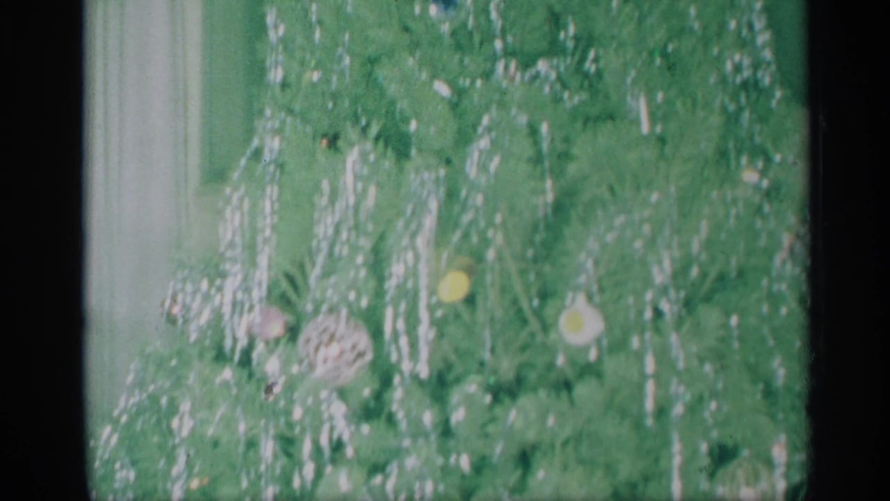 Decorated Christmas tree branches fill the frame, with white tinsel and round ornaments visible in soft 8mm color.