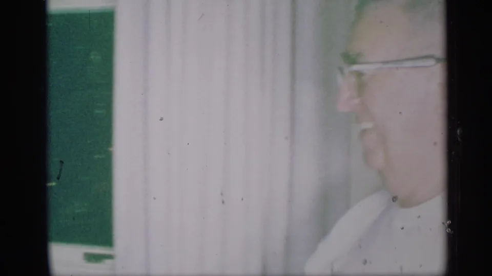 1959 New York home-movie frame showing an adult man in glasses in profile near pale curtains inside a domestic room.