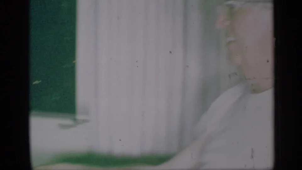 1959 New York home-movie frame showing an elderly man in glasses and a white shirt, softly framed near curtains.