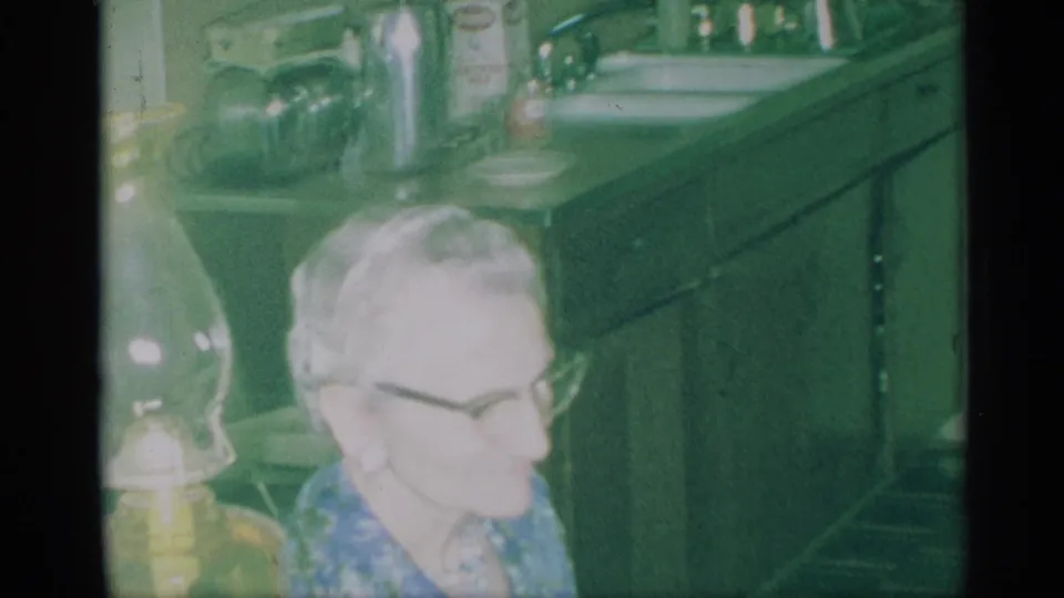 1959 New York home-movie frame showing an older woman with glasses near a kitchen counter, cabinets, and household appliances.