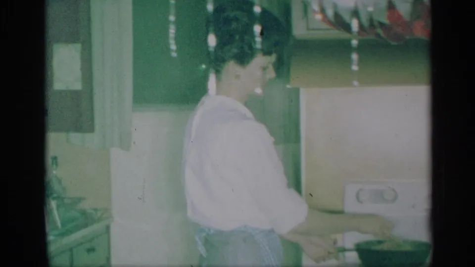 1959 New York home-movie frame showing a woman in a light blouse working at a kitchen counter inside a decorated home.