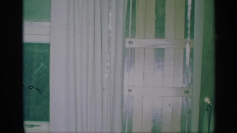 1959 New York home-movie frame showing white curtains, green blinds, and part of a window inside a private room.